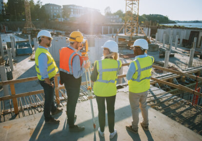 Construction Worker Using Theodolite Surveying Optical Instrument for Measuring Angles in Horizontal and Vertical Planes on Construction Site. Engineers and Architect Discuss Plans Next to Surveyor.